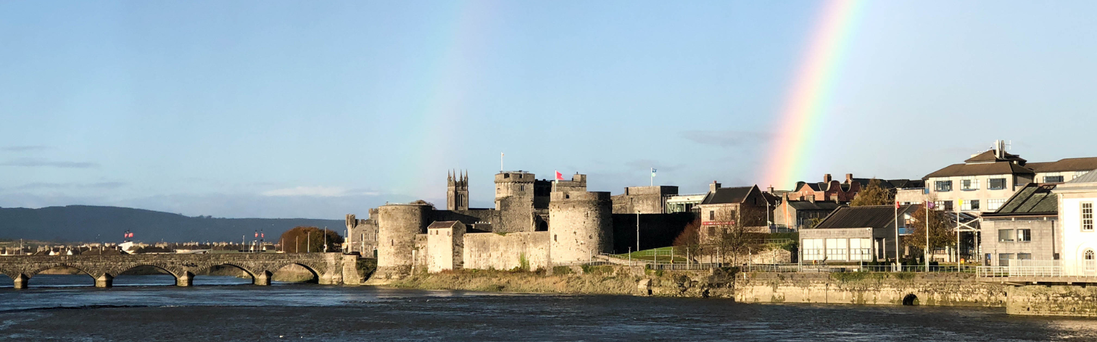 Two rainbows emerge behind a dark, stone castle with various circular turrets, sitting on the edge of a river with a bridge in the distance. 